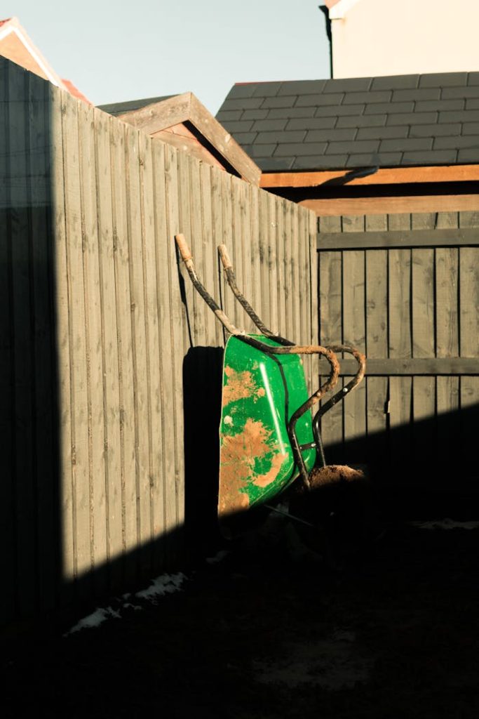 wheelbarrow-in-yard-11408153 A rustic green wheelbarrow leaning against a wooden fence in a sunny village yard scene.